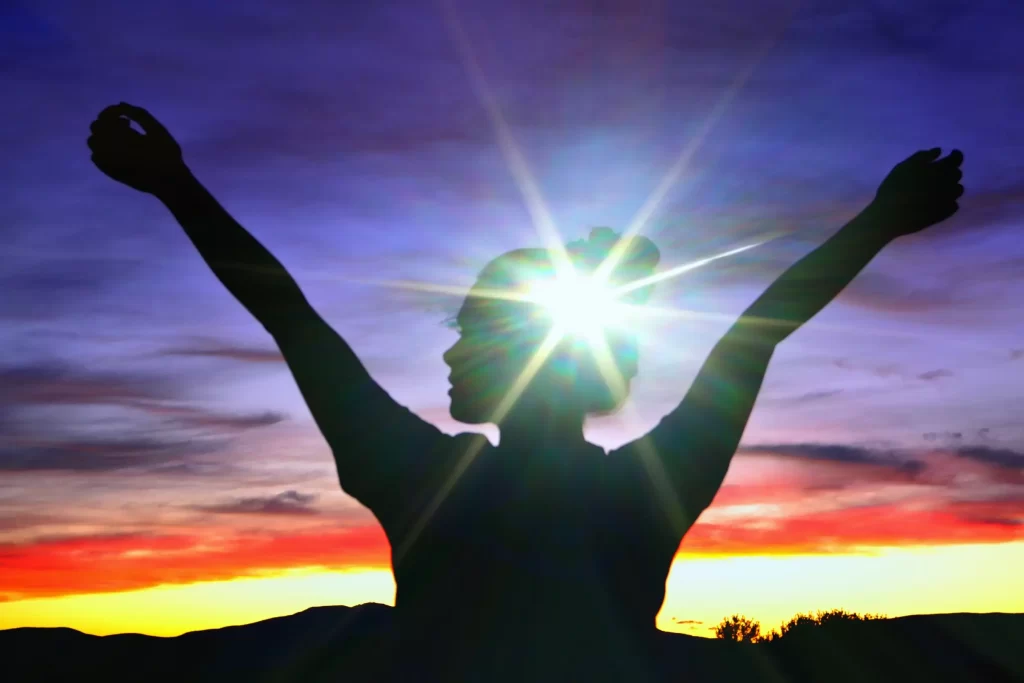 A silhouette of a woman raising her hands with sun rays reflecting on her.